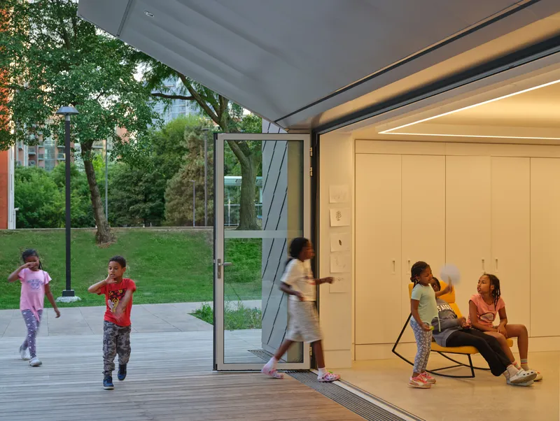 Open glass doors connect the interior room to children playing on the exterior deck at dusk