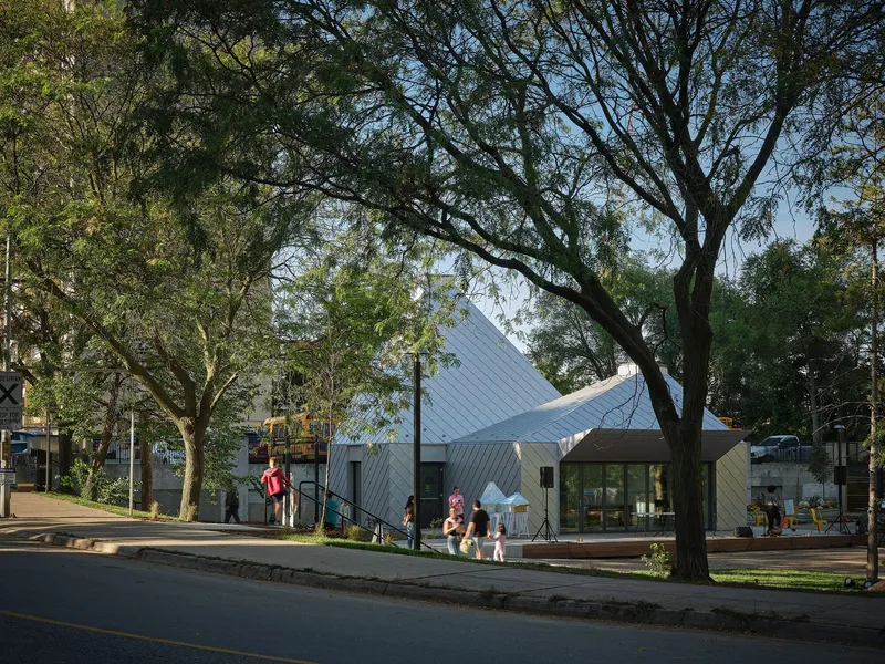 Street view of the white metal tiled pavilion beneath the canopy of park trees in late afternoon