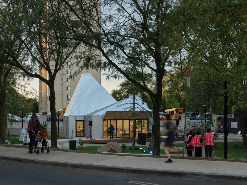 Pavilion with twin pitched roofs and open glass facade framed by pedestrians and park trees at dusk