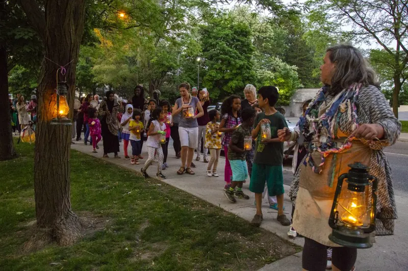 Evening procession along tree-lined sidewalk with participants carrying lanterns at dusk