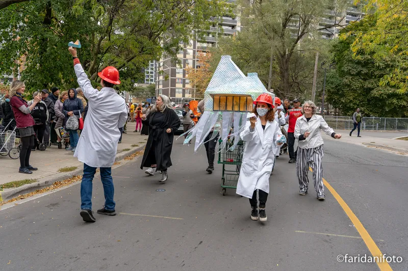 Street parade with costumed participants wheeling paper model with checkered pyramidal roof through autumn leaves
