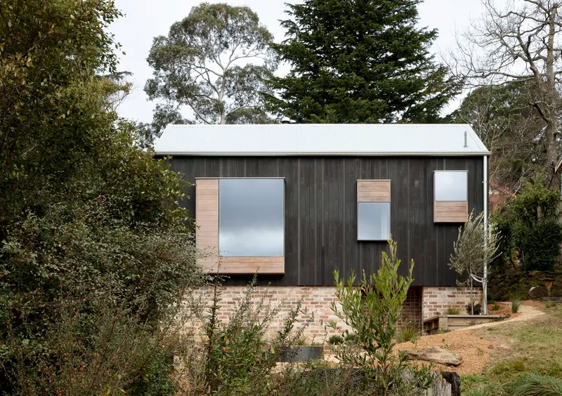 Side view of the dark timber clad volume on brick plinth set into a hillside with eucalyptus trees