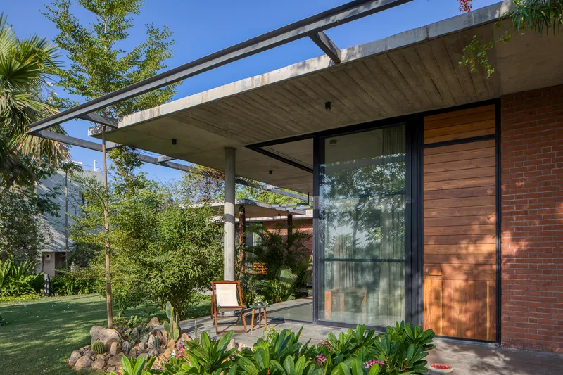 Covered terrace with board-formed concrete ceiling and glass walls framing a planted courtyard with dappled sunlight