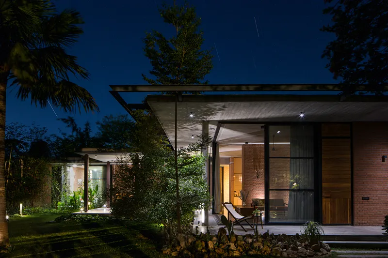 Garden view of the flat-roofed pavilion with floor-to-ceiling glazing and exposed concrete soffit at dusk