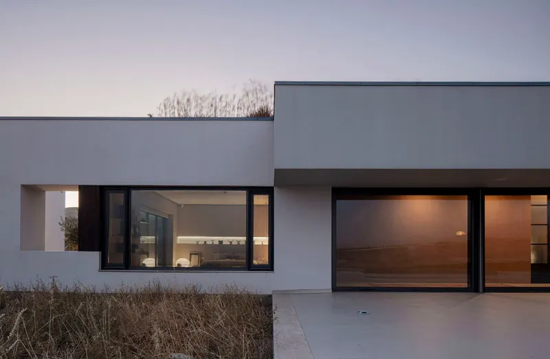 Illuminated interior visible through black-framed glazing at dusk with parched grasses in foreground