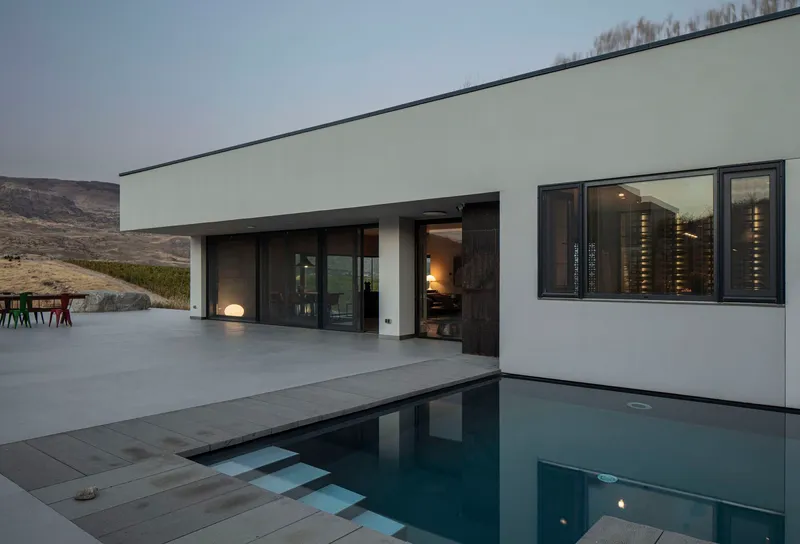 Pool deck at dusk with still water reflecting the interior lights through sliding glass doors