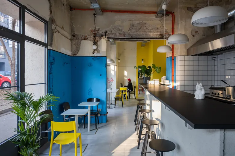 Cafe interior with exposed concrete ceiling, blue painted walls, white tile kitchen counter and yellow seating