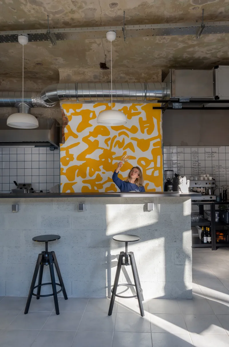 Bar counter with metal stools in streaming sunlight beneath yellow patterned wall panel and exposed ductwork