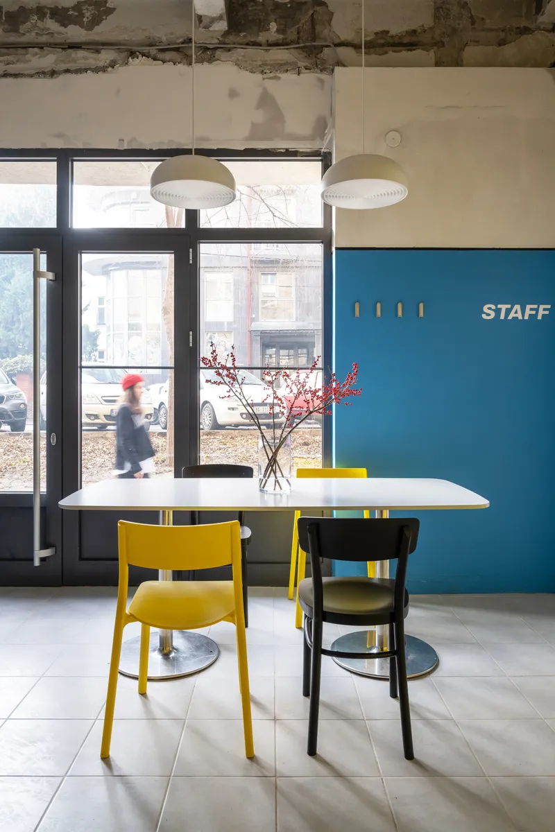 Dining area with white table, yellow and black chairs, and a person walking past the street-facing glazing