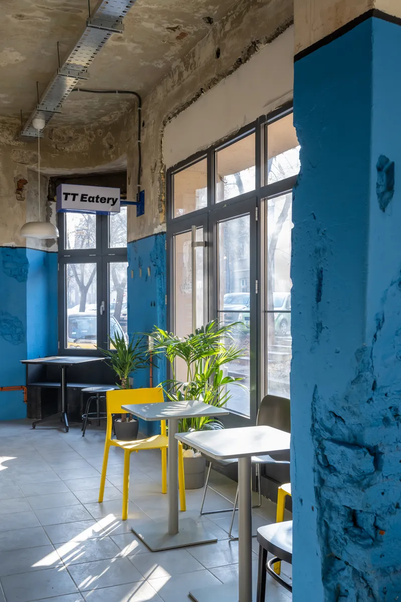 Seating area with small tables, potted plants, and blue-painted columns near floor-to-ceiling windows