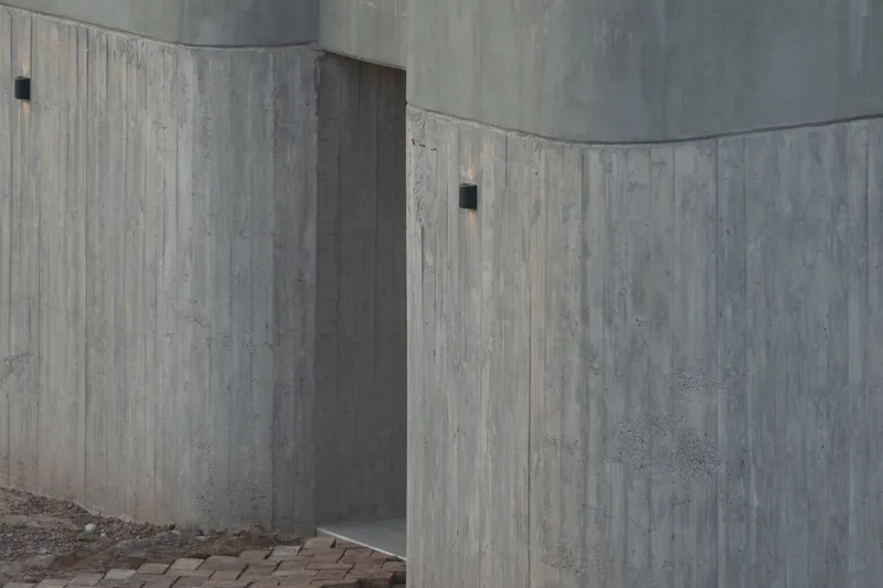 Detail of interlocking board-formed concrete walls with small wall-mounted fixtures at the entrance