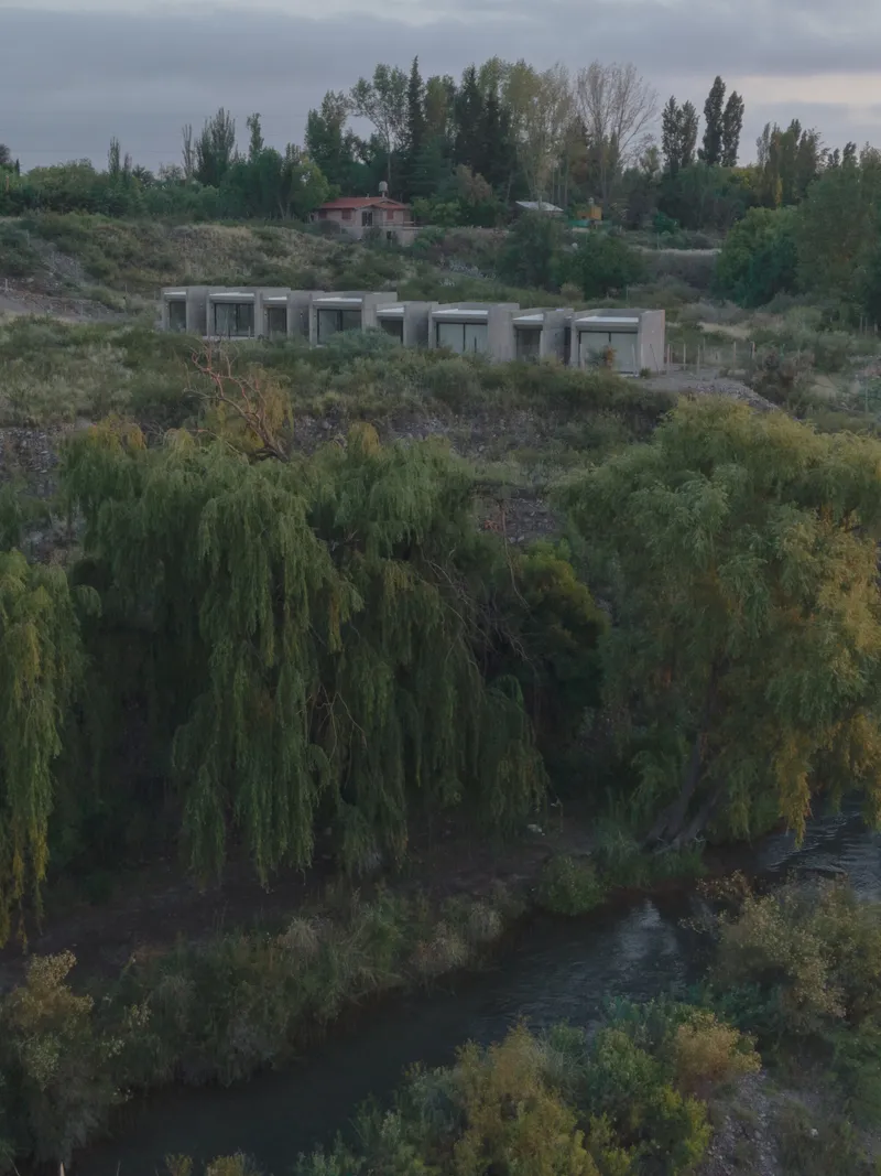 Long concrete volume set into a terraced hillside above a river with willow trees at dusk