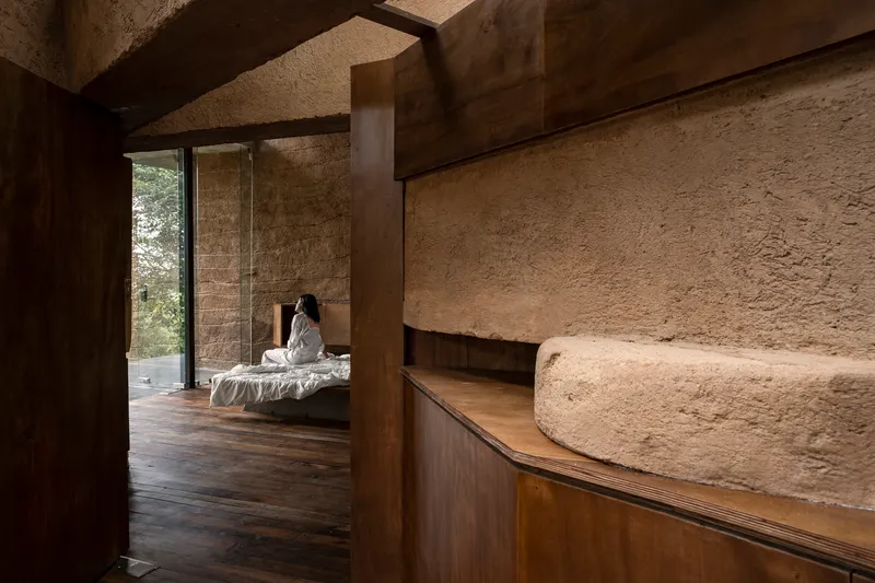 Curved wooden millwork meeting textured earthen walls with a figure seated on a bed beyond