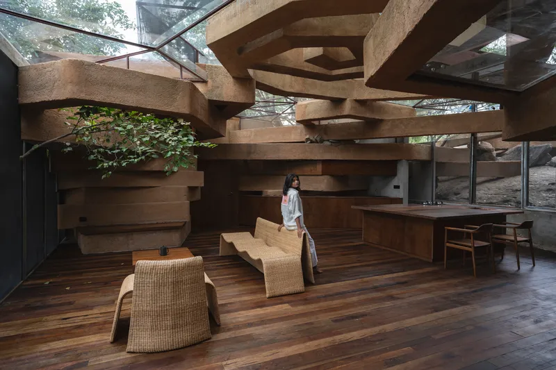 Interior living space with stacked rammed earth beams overhead and a woman standing among woven seating