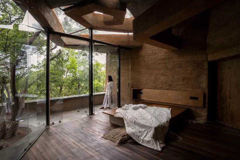 Bedroom with floor-to-ceiling glass walls opening to trees and the adjacent rammed earth wall