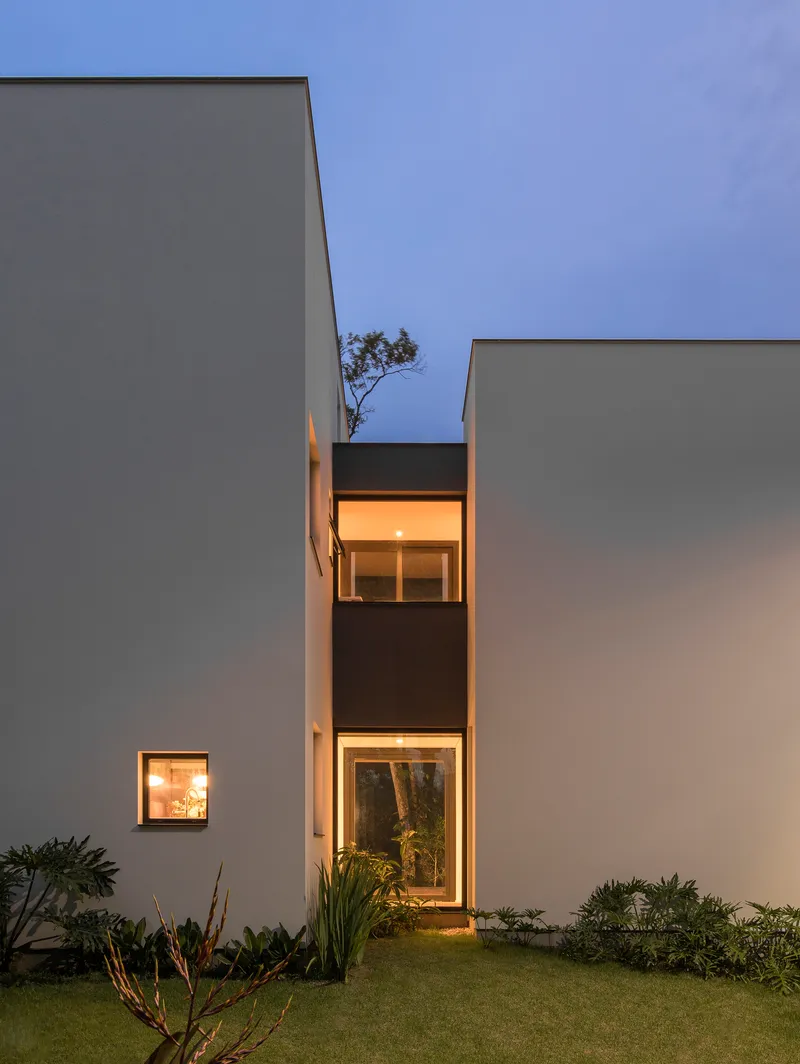 Illuminated windows recessed into white volumes framing a small tree between planted beds at twilight