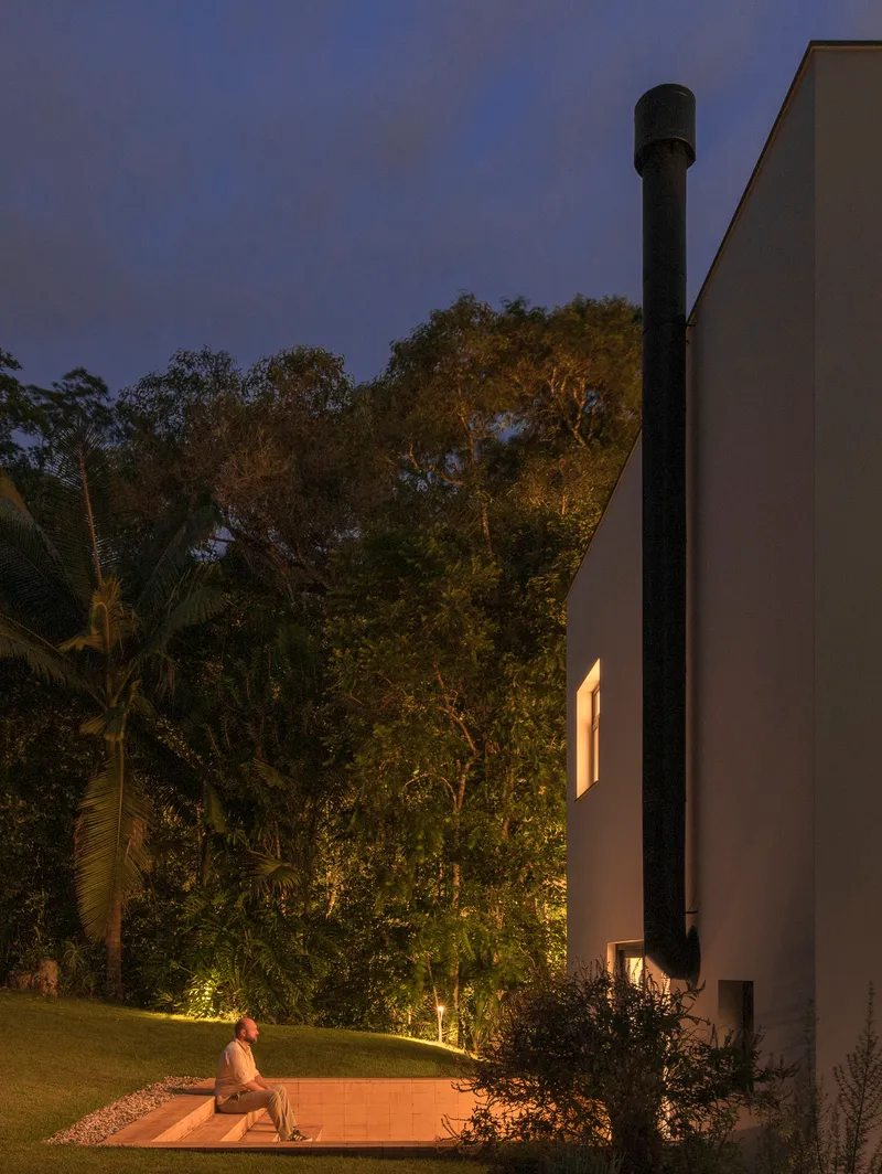 Black cylindrical flue rising against the white facade with a figure seated on timber deck at dusk