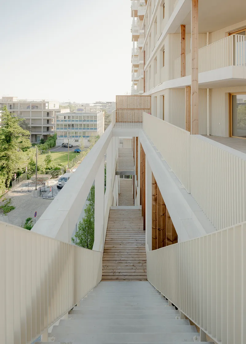 Timber deck walkway framed by vertical corrugated panels looking toward neighboring buildings and trees