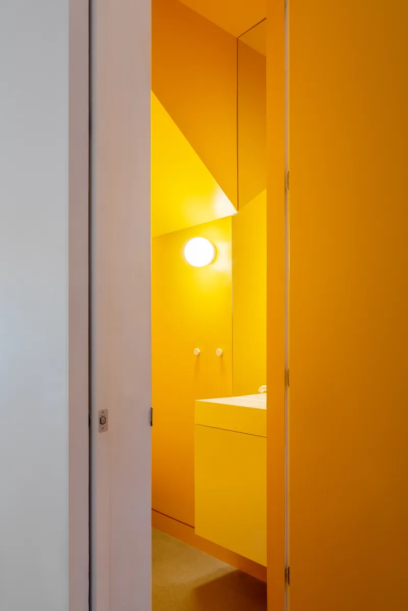 View through a narrow doorway into a fully tiled bathroom with yellow surfaces and circular wall sconce
