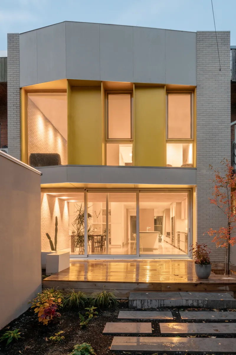Rear garden facade with warm-lit glazing and timber deck stepping down to planted beds at dusk
