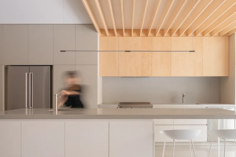 Kitchen counter with blurred figure moving past stainless steel refrigerator and pale wood cabinetry