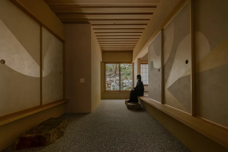 Narrow corridor with exposed timber ceiling beams and a seated figure facing a garden window