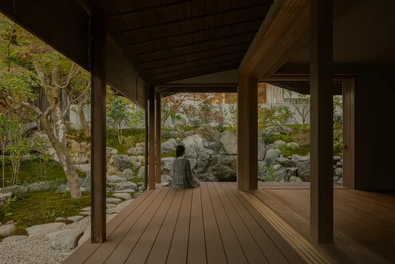Covered timber deck with a seated figure facing a moss and stone garden in autumn light