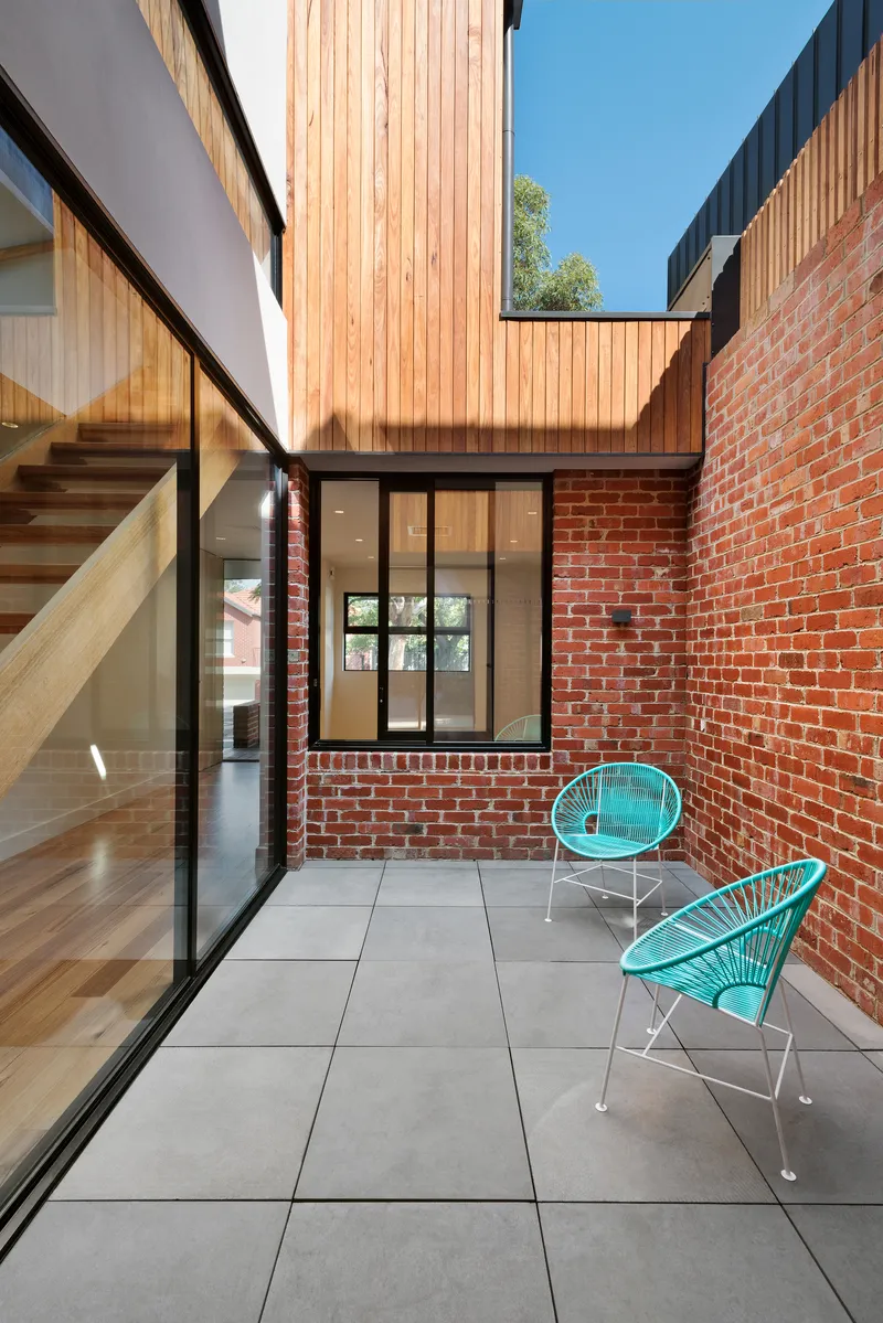 Internal courtyard with large-format grey pavers, red brick walls, and two turquoise wire chairs under blue sky