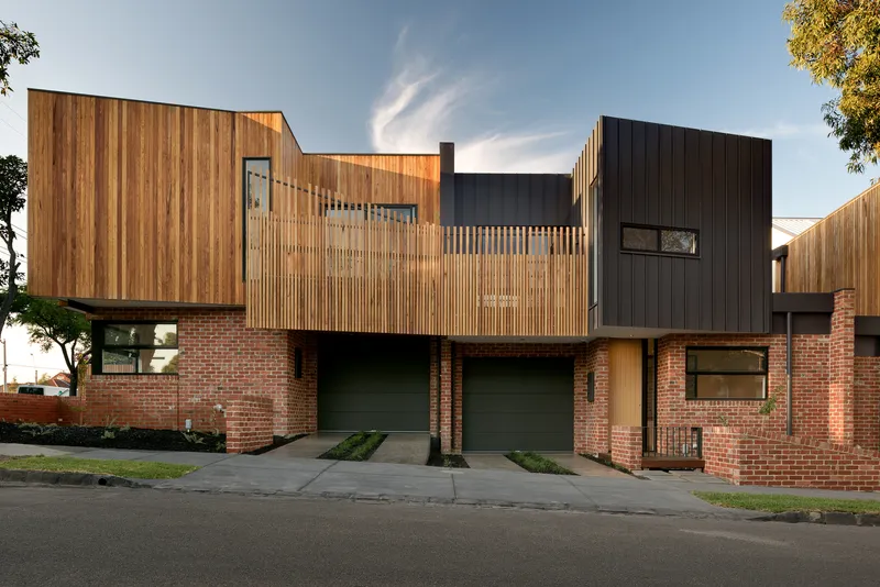 Front elevation at dusk showing the layered facade of brick base, timber screens, and dark metal volumes with garage doors below