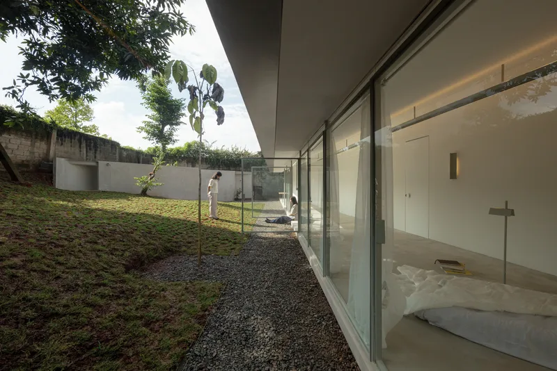 Glass-walled bedroom opening to a courtyard where two figures stand beside a young tree