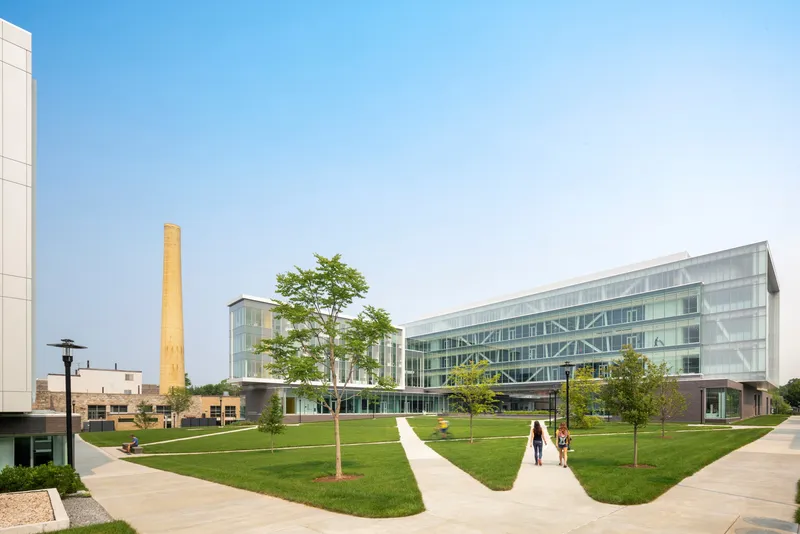 Campus green with diagonal pathways connecting glazed volumes and brick smokestack under clear sky