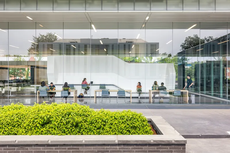 Ground-level view through floor-to-ceiling glass showing a study area with planted hedge bed in foreground