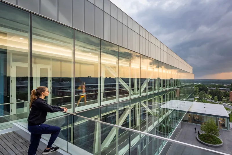 Upper-level terrace with glass balustrade where a person sits near the illuminated facade at dusk