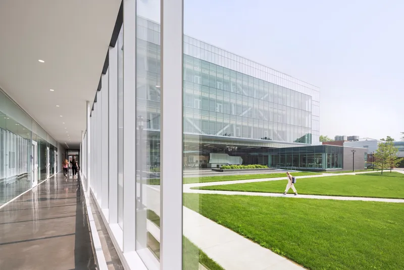 Interior corridor with floor-to-ceiling glazing overlooking the lawn where a figure walks across the courtyard