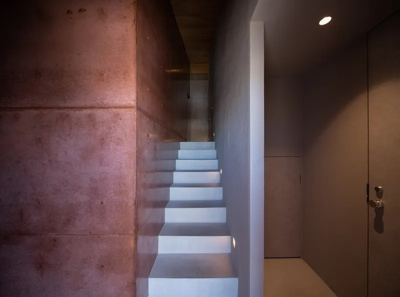 Concrete staircase rising between exposed aggregate walls and white plaster surfaces under recessed lighting