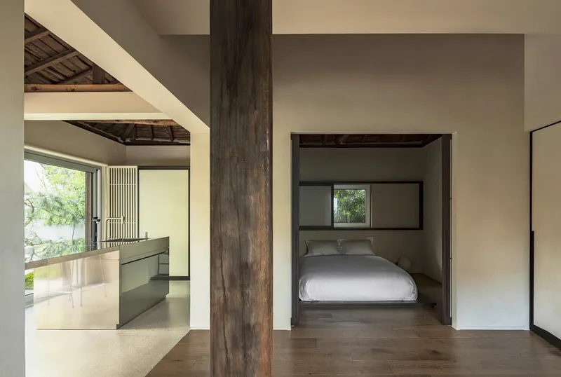 View through a dark timber column toward a bedroom alcove and kitchen with exposed roof trusses