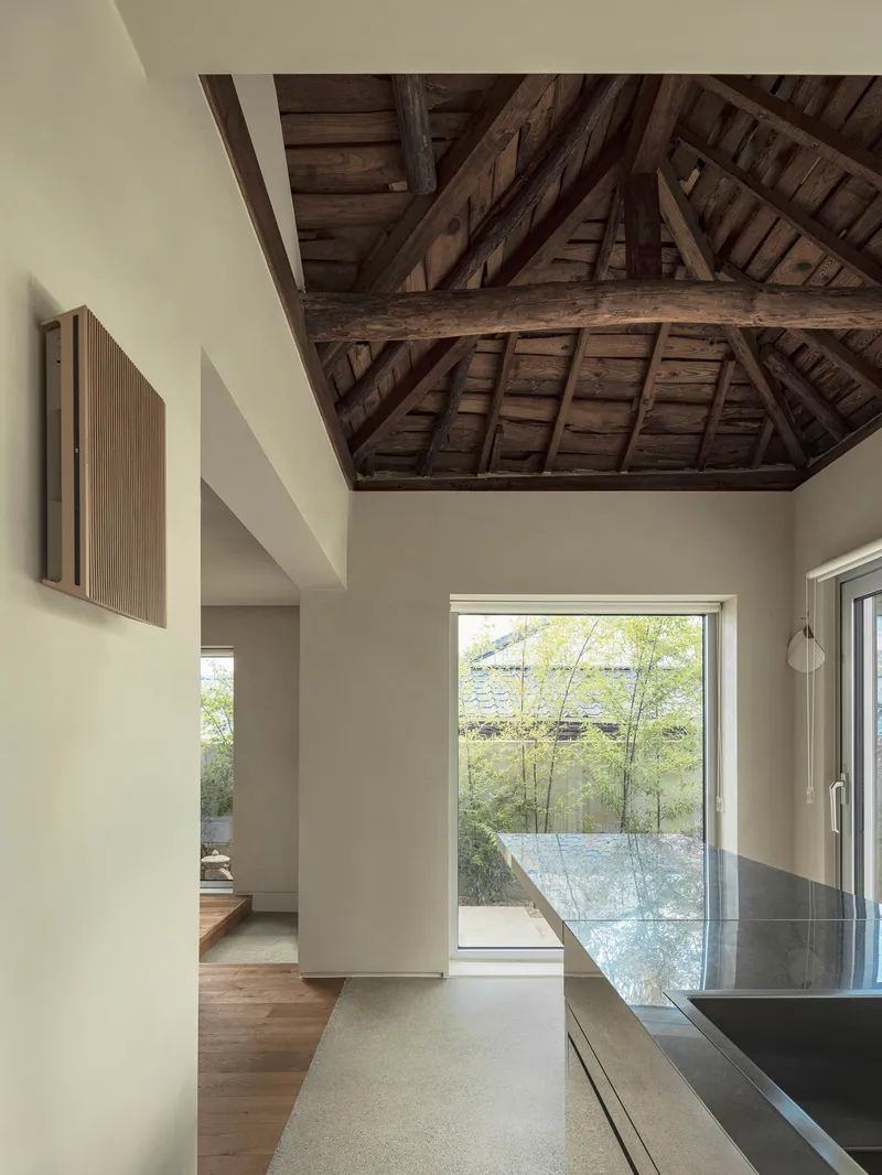 Kitchen interior with exposed timber roof structure and window framing a gravel courtyard with bamboo