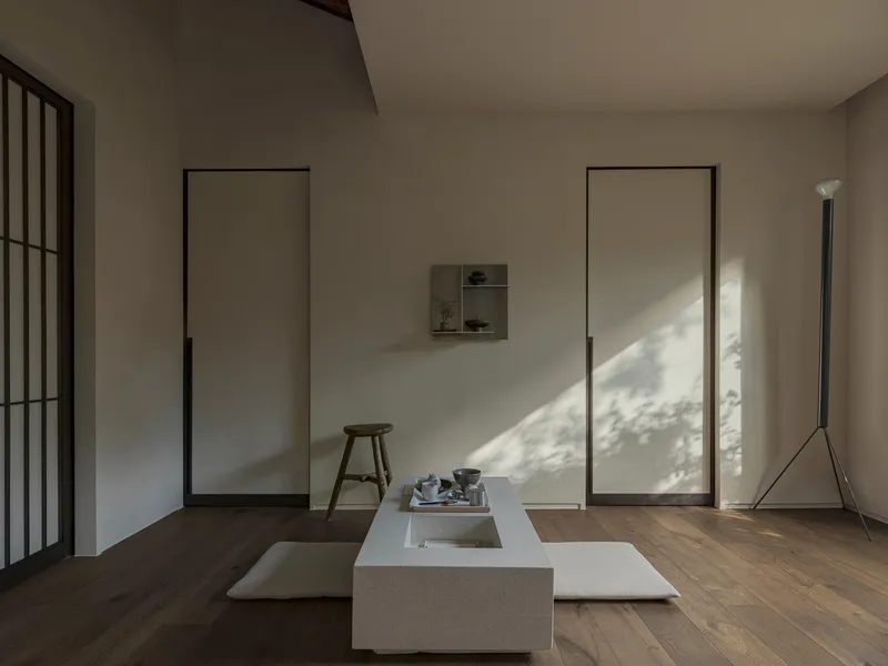 Minimalist interior with flush panel doors, low stone platform and dappled sunlight across oak floors