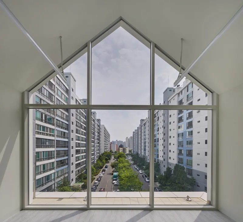 Interior view through gabled window framing tree-lined street and apartment blocks beyond