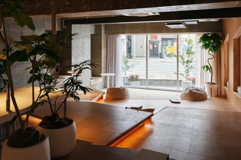 Interior lounge area with exposed concrete ceiling beams, potted plants, and a large glazed storefront opening to the street