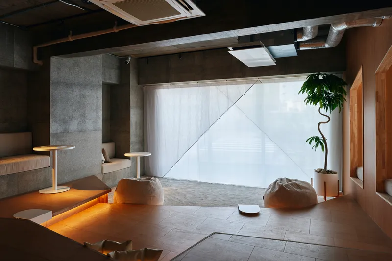 Living space with concrete columns, sheer curtains filtering daylight, and floor cushions on grey tile flooring