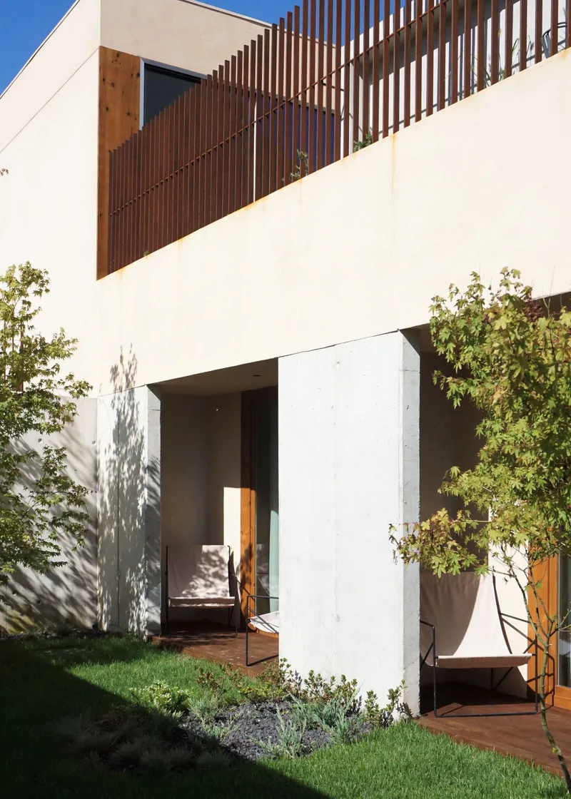 White stucco facade with timber screen balcony and dappled tree shadows across the entrance threshold below