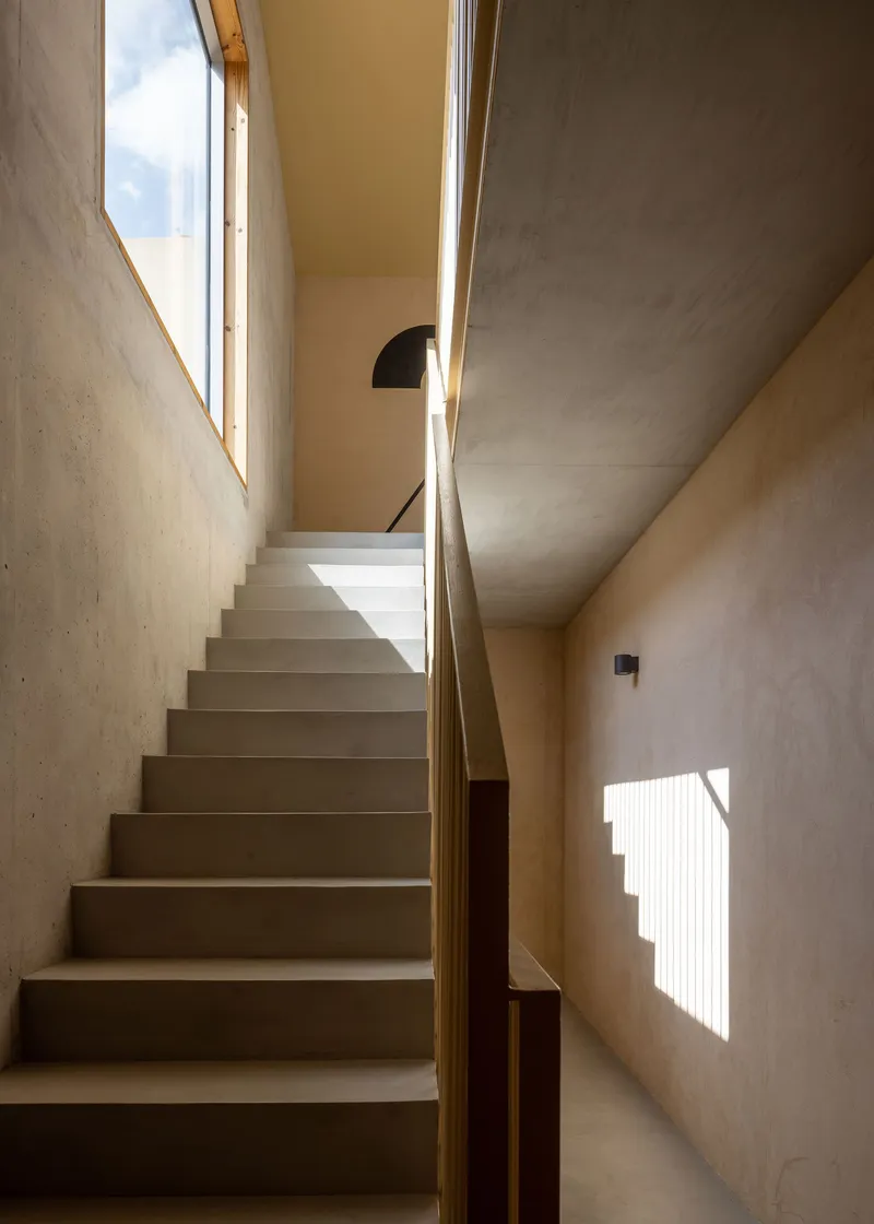 Concrete staircase ascending through a skylit volume with plywood walls and shadow patterns