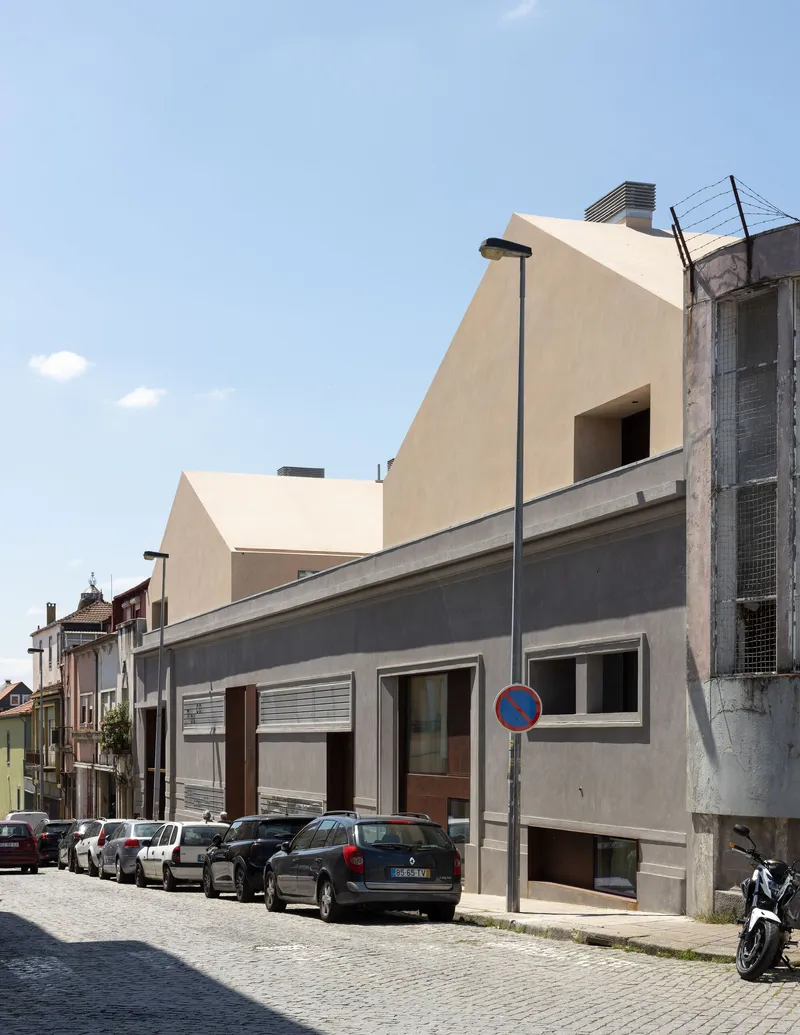 Street facade with cream volumes rising above a grey rendered base and parked cars along the cobblestone