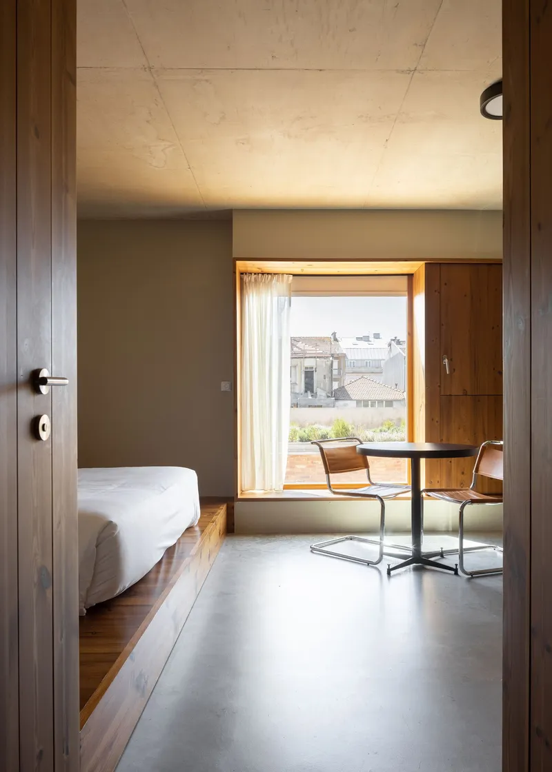 View through timber doorway into bedroom with concrete ceiling and window seat bathed in daylight
