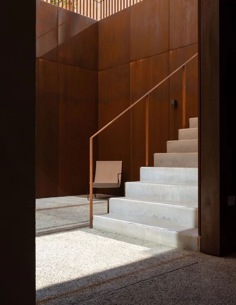 Concrete stairs with copper handrail against weathered steel panel wall in a sunlit courtyard