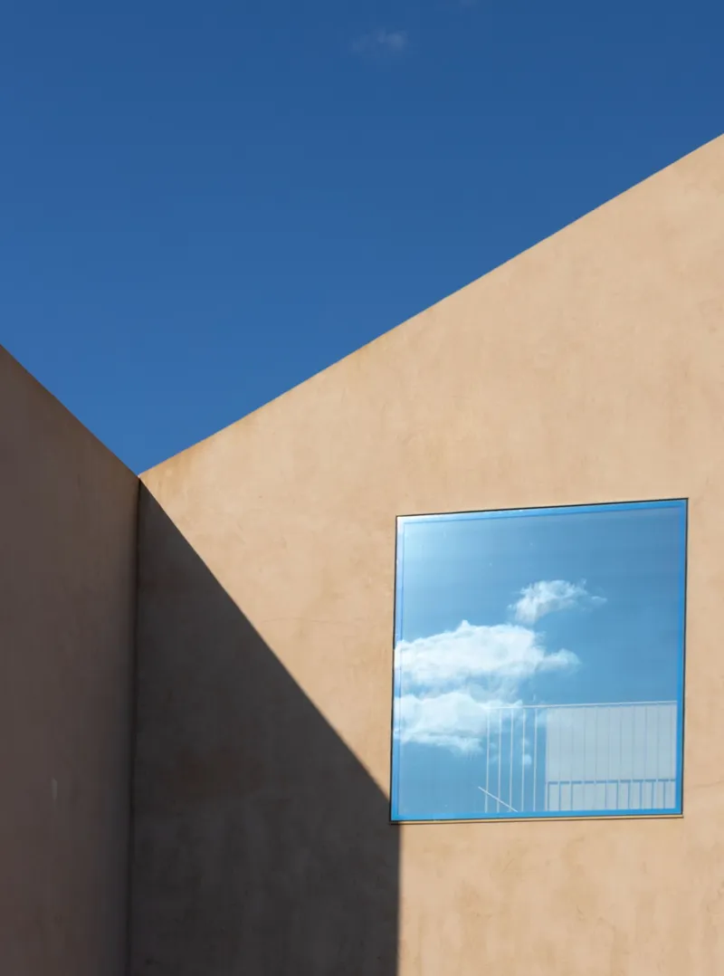Square window framing clouds and a distant facade within a peach-toned stucco wall under blue sky