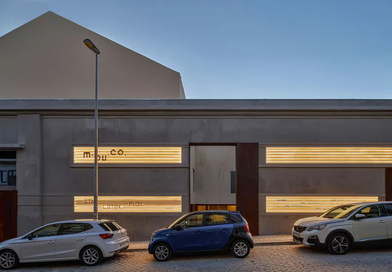Street facade with horizontal lit openings behind concrete panels at dusk, parked cars in foreground