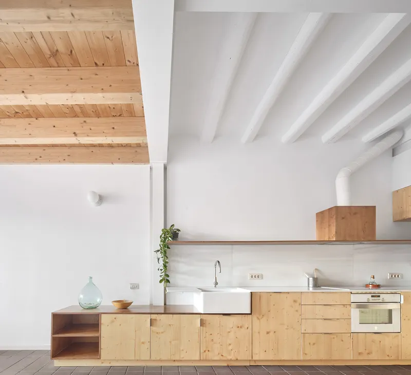 Kitchen with light pine cabinetry beneath a double-height space with exposed timber plank ceiling