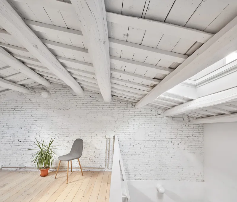 Upper floor room with whitewashed brick wall, exposed timber beams and a potted plant beside a chair